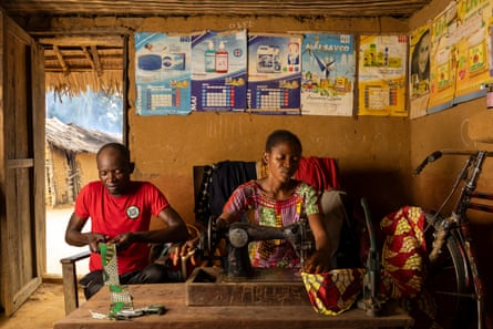 A man holding a piece of cloth sits next to a woman with a sewing maching in a small building