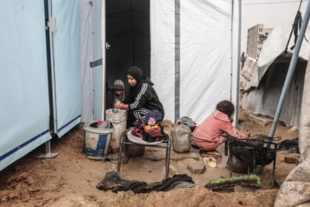 Women and children crouch in the entrance of a tarpaulin shelter, washing clothes in buckets, the ground around them muddy and with deep puddles.