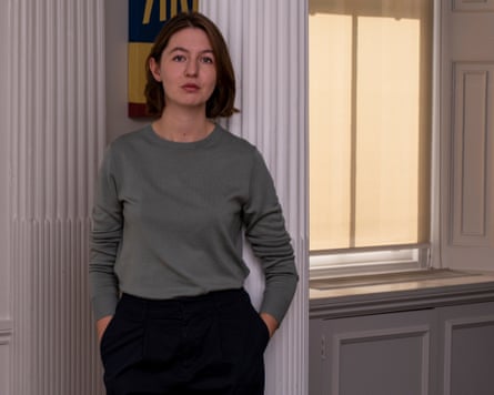 Sally Rooney stands in a white-painted, wooden panelled room near a window. She has straight, light brown collar-length hair and wears a pale green jumper and dark trousers. She is in her late 20s in this 2018 picture.