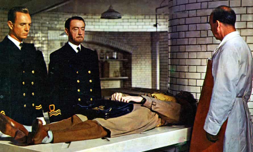 Two men in military uniform and a doctor in a white coat look at the body of a man on a slab