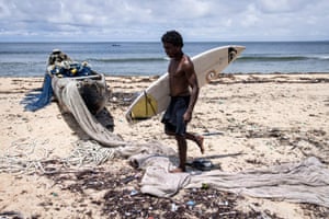 Samuel carries his surfboard past a pirogue on his way to help pull a fishing net in and then surf in Robertsport