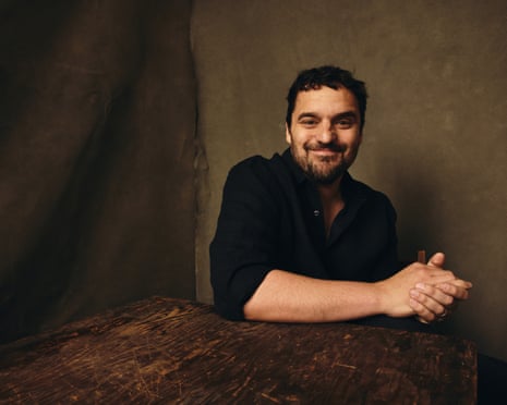 Jake Johnson in a black shirt sits at a dark wooden table, smiling at the camera