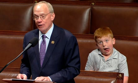 A boy makes a silly face by sticking his tongue out and opening his eyes wide as he sits next to a politician in the House of Representatives
