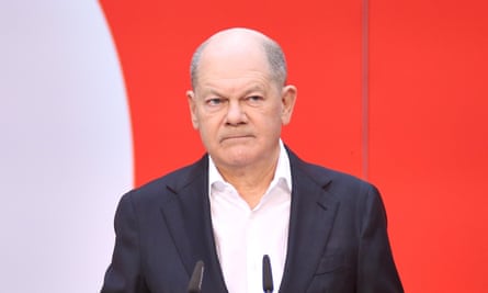 Olaf Scholz looking annoyed in a dark blazer, standing in front of a red and white background