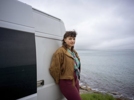 A young woman leans against a white van with the sea in the background