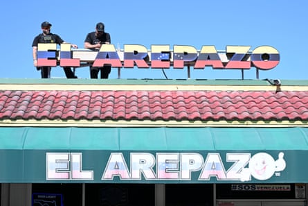 Two men on top of a red-tiled roof leaning on sign that says El Arepazo.