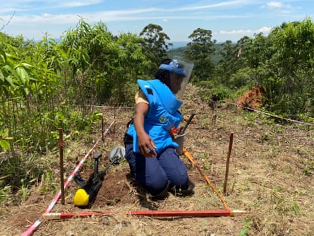 An African woman wearing protective clothing kneels in a field next to crops with a metal detector beside her