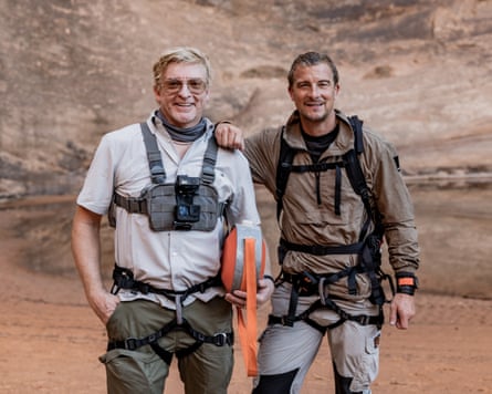 Rhys Darby and Bear Grylls, wearing hiking gear and harnesses, stand side by side in a rocky desert canyon.