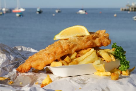 Close-up image of cardboard takeaway tray of fish and chips with mushy pea fritter, slice of lemon, parsley, wrapped in paper.