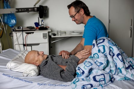 Man in a T-shirt and glasses smiles as he puts his hand on the hand of a man in a fleece, smiling, who is lying in a hospital bed under a duvet