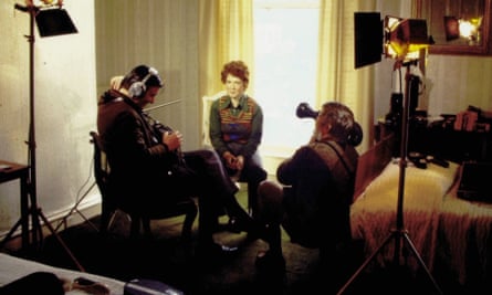 A kneeling cameraman films director Zwy Aldouby, wearing headphones and seated opposite a woman with short hair wearing a vest over a long-collared shirt, in a hotel room set up with spotlights