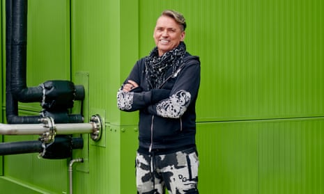 Dale Vince standing outside a corrugated iron industrial facility painted green. He is wearing black and white cargo pants and a sweatshirt with skulls on the elbows. His hair is swept to one side.