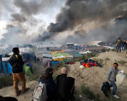 Migrants watch thick black smoke rise over burning shelters in the Calais “Jungle” camp