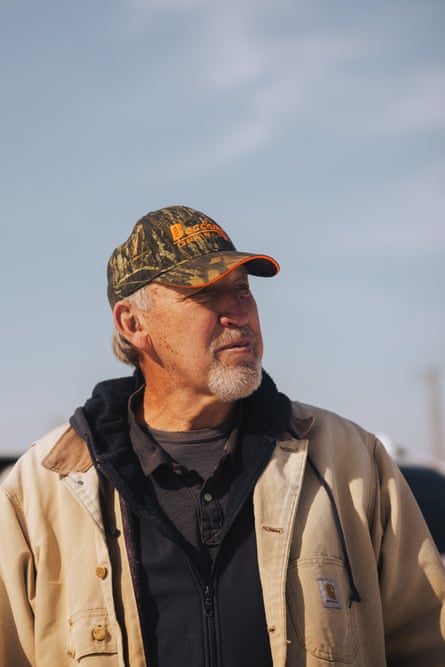 a man in a brown jacket and camo hat stands for a portrait against the sky as a backdrop