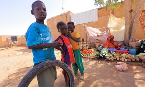 Three little boys, one with a tyre used as a toy, in a market place with a vegetable seller in the background