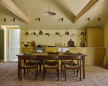A large kitchen painted in pale yellows, with a brick floor, a wooden dining table and pale beams