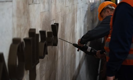 Two metro workers wearing orange vests and orange hard hats install new letters on the off-white wall of of a metro station in Kyiv, Ukraine. 
