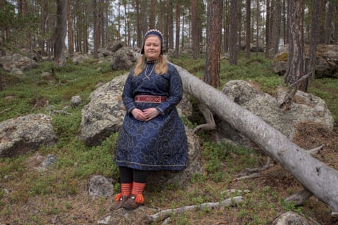 A blond woman in a traditional dress, woven hat and belt, and embroidered boots, sitting on a rock in a forest