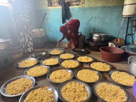 A woman next to vats puts rice on a dozen metal platters on the floor.