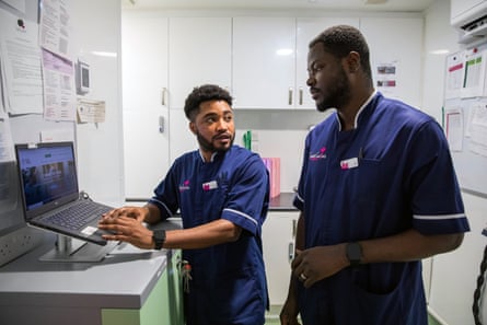 Two men in nursing tunic tops converse next to a laptop.