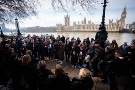 People gather in a group on the path with the River Thames and Houses of Parliament seen in the background