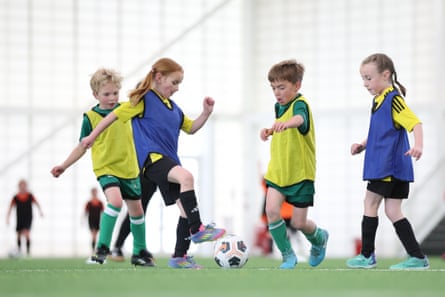 A group of four children, two girls and two boys, play a game of 3-v-3 football on an indoor pitch.