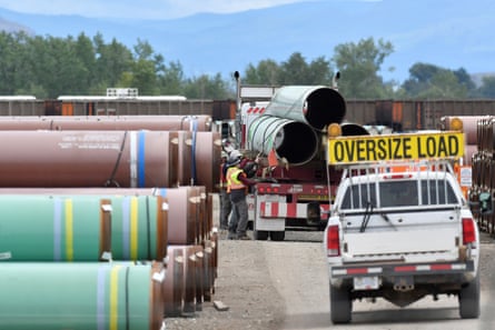 At a pipe yard, a white pickup truck has a yellow and black sign reading ‘Oversize Load.’