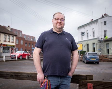 Chris Taylor stands on a street in Rochford in glasses and a navy polo shirt, with houses and cars behind him