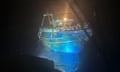 The Adriana, an ageing fishing trawler, is seen illuminated in the dark at a port with people crammed onto its decks and even onto the roof of its cabin. It is painted blue with large patches of rust on its hull.