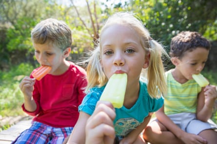 three young children in garden eating ice lollies; a girl in the centre is leaning to the camera holding a yellow lolly in front of her mouth while two boys sit to either side eating their lollies