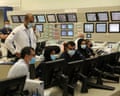 Workers wearing face masks sit at computer workstations in a control room with monitors and control panels
