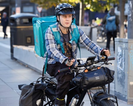 Man in checked shirt, black cycle helmet and blue insulated backpack rides his bicycle.