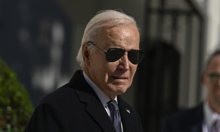 Elderly white man with white hair to his neck wearing a black trench coat and aviator sunglasses.