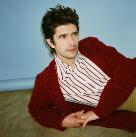 Actor Ben Whishaw in dark red cardigan an d striped shirt, lying on wooden floor, against blue background, January 2022