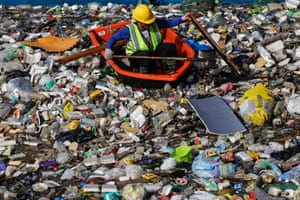 A man in a hard hat and hi-vis jacket in a creek full of rubbish in a small red rowing boat
