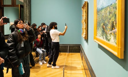 A bearded man in a white T-shirt holds up a phone in front of a painting, centre, while others look at their screens.