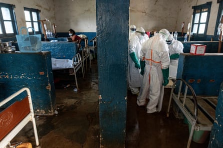 People wearing PPE work in a run-down ward with metal beds.