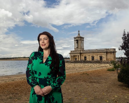 A dark-haired woman in a green and black patterned dress poses alongside a lake with a grand building in the distance.