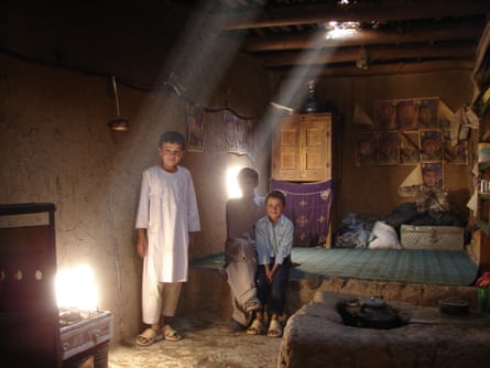 Three children stand in a mud-walled room with a shaft of light coming from a hole in the ceiling