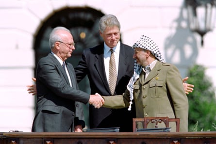 A taller white man with gray hair in a suit stands with his arms outstretched toward two shorter men who are shaking hands. The man on the left is white, mostly bald and wearing on a suit. The man on the right is Arab, with a beard, a keffiyeh wrapped around his head and hanging down one shoulder, and an olive-colored jacket.