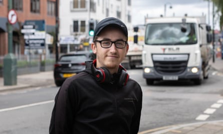 Josh Frost, in a baseball cap and with glasses and headphones resting on his shoulders, smiles slightly with the road and a lorry behind him