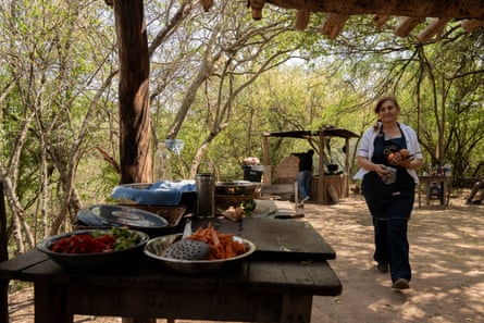 A woman carrying vegetables walks towards a table covered in bowls full of food outside under a shelter surrounded by trees