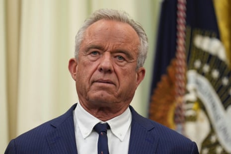 Robert F. Kennedy Jr. stands with a serious expression in the Oval Office, flags blurred behind him