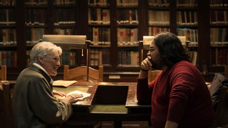 Glenn Close and Carlos Gonzalez sit across a desk talking in a library lined with bookshelves