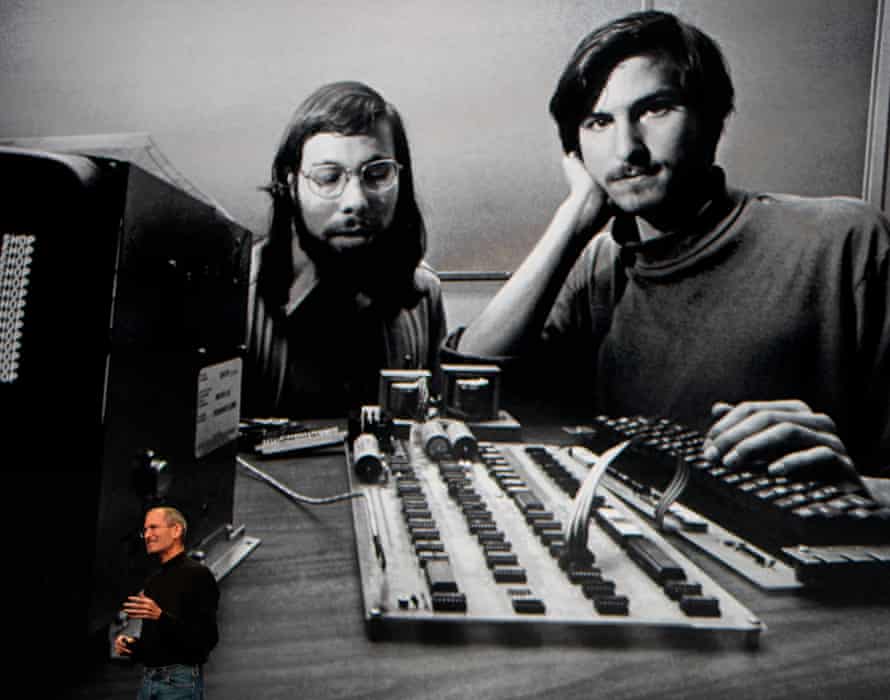Steve Jobs stands beneath a black and white photograph of him and Steve Wozniak with an early computer