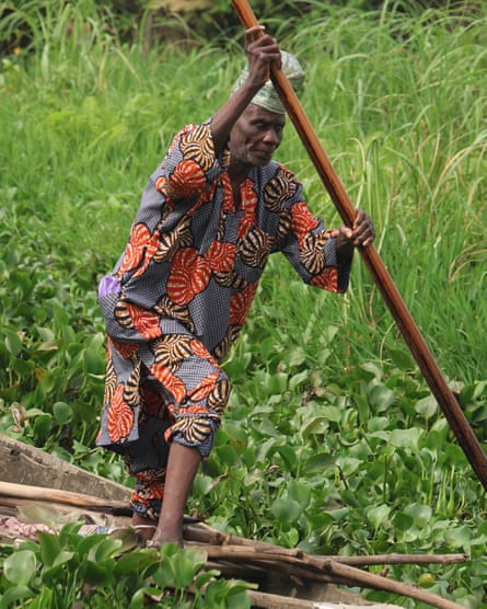‘The water is no longer our friend’: how dredging is pushing Lagos Lagoon towards ecosystem collapse – photo essay An older man wearing a colourful robe uses a pole to push a boat through water plants.