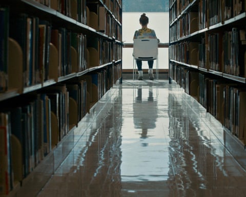 A young woman sits at a table in a library at the end of two rows of books