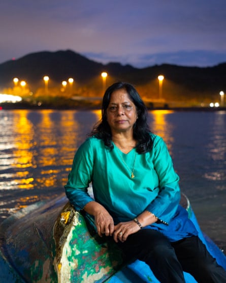 A woman in a green and blue top and dark trousers reclines upon a rock by water, with the lights of Hong Kong harbour behind her