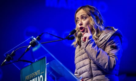 Giorgia Meloni speaks at a rally for regional elections in Abruzzo, 5 March 2024: she is a woman in her mid-to-late 40s with long blond hair worn loose and lightly waved, and is seen against a deep blue backdrop; she is wearing a light brown padded coat