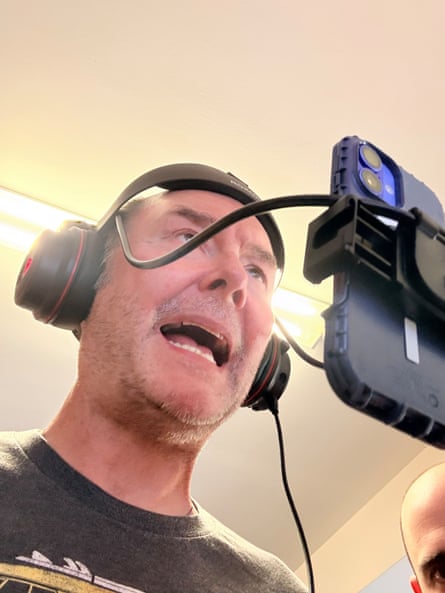 Dominik Diamond wears headphones and speaks animatedly into a smartphone on a selfie rig